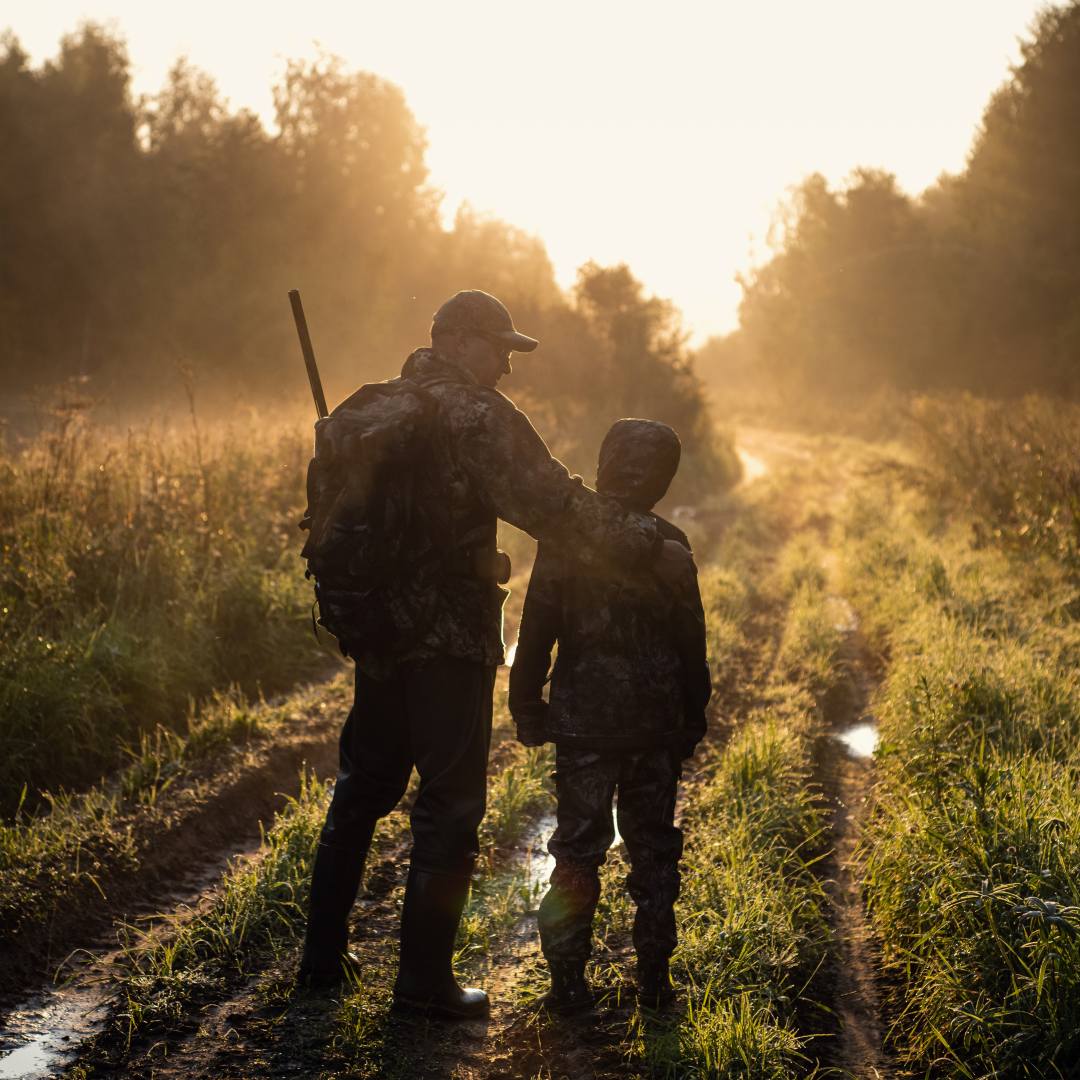 ALT: An adult and child walking together in a field with a sunset in the background geared up for a hunting trip.